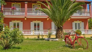 A two-story building with a red facade, white balconies, and a palm tree in the front yard. - Spacious first floor apartment, Family-Friendly Stay Near the Beach (Corfu)