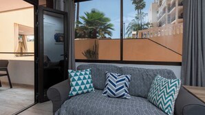 Un salon moderne avec un canapé, des coussins à motifs, une porte-fenêtre coulissante et une vue sur des palmiers et des immeubles.