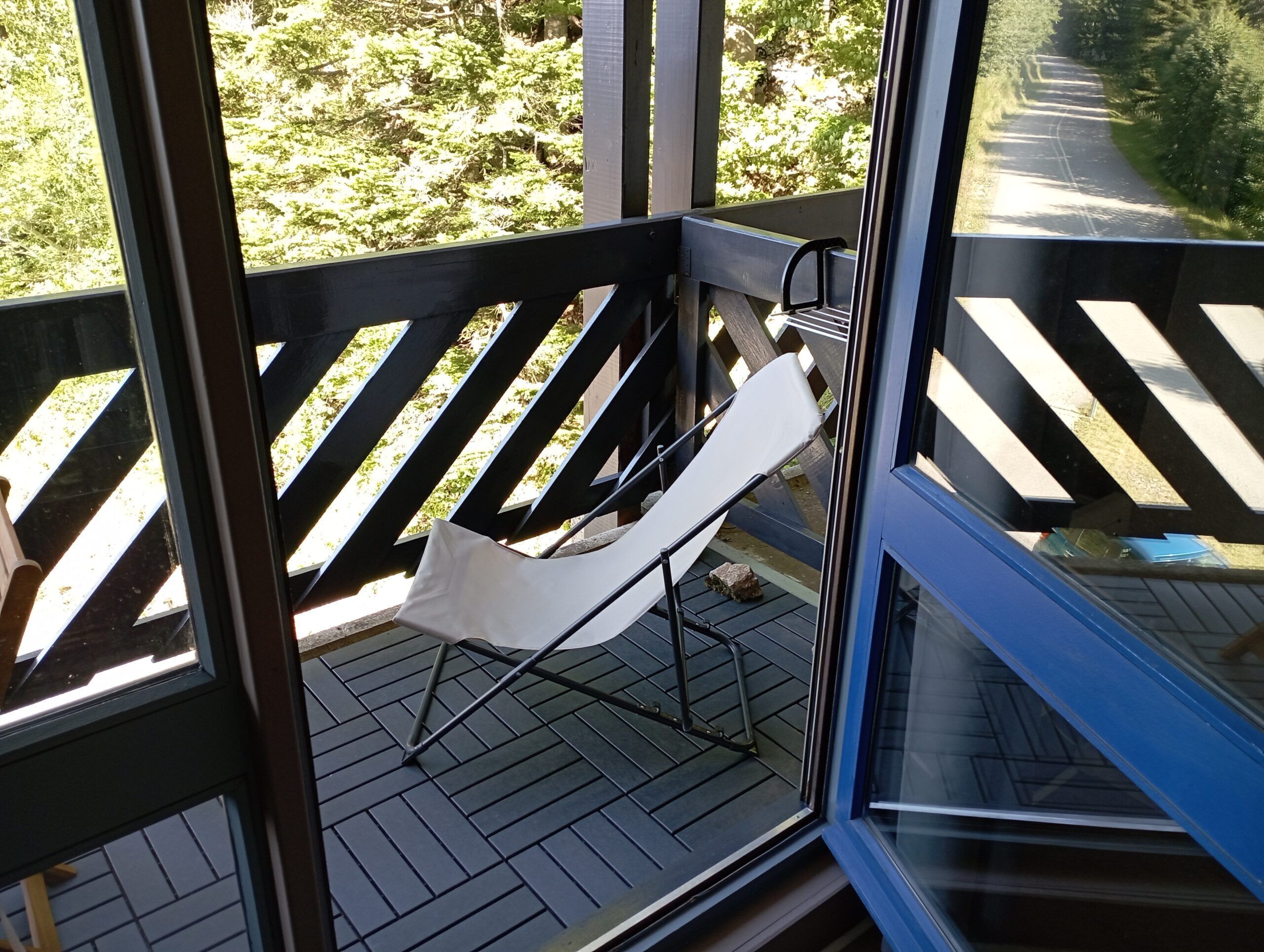 A balcony with a white hammock chair, wooden floor, and a view of a road and greenery.