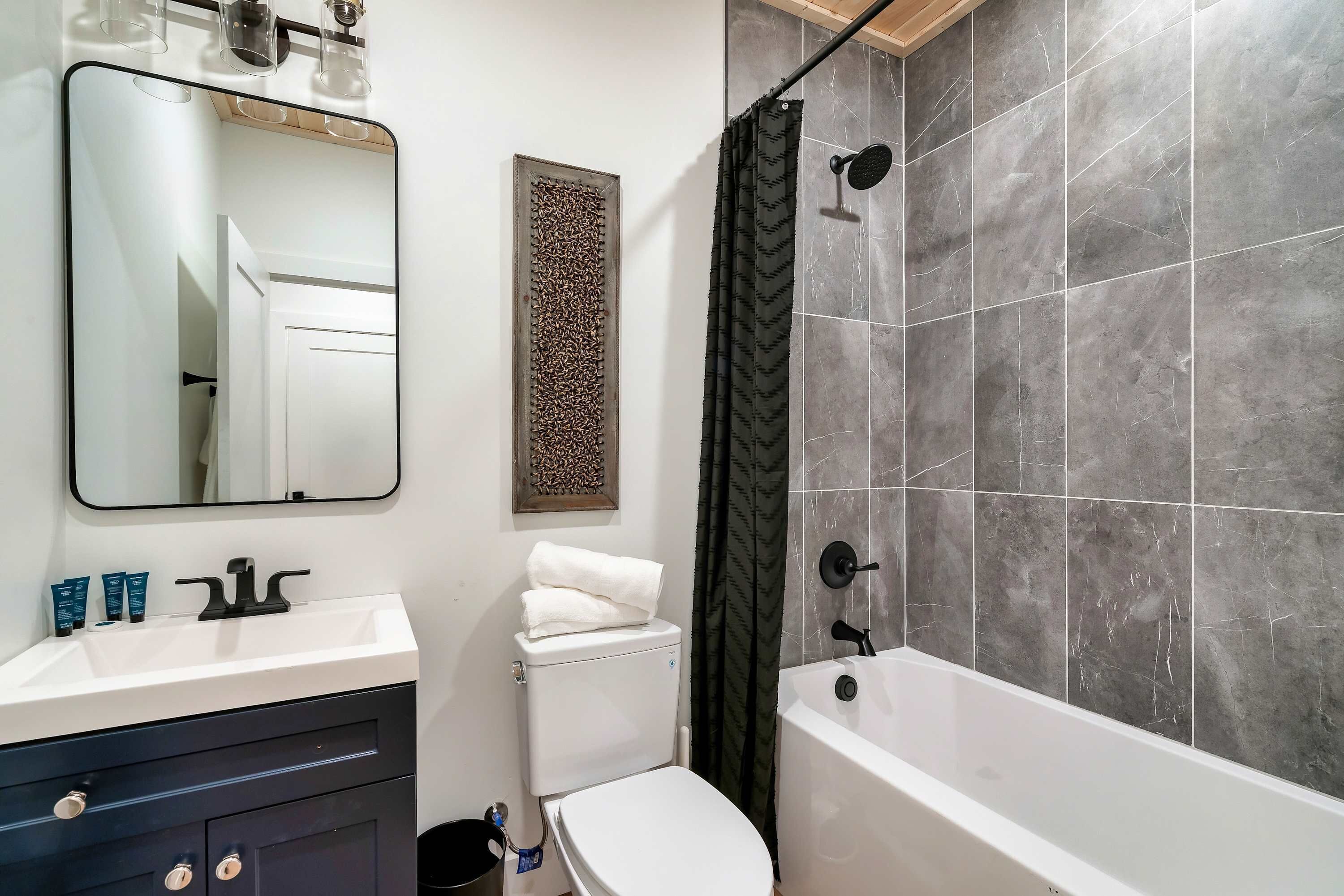 A modern bathroom with a white sink, dark cabinets, a white toilet, and a bathtub with grey tiles.