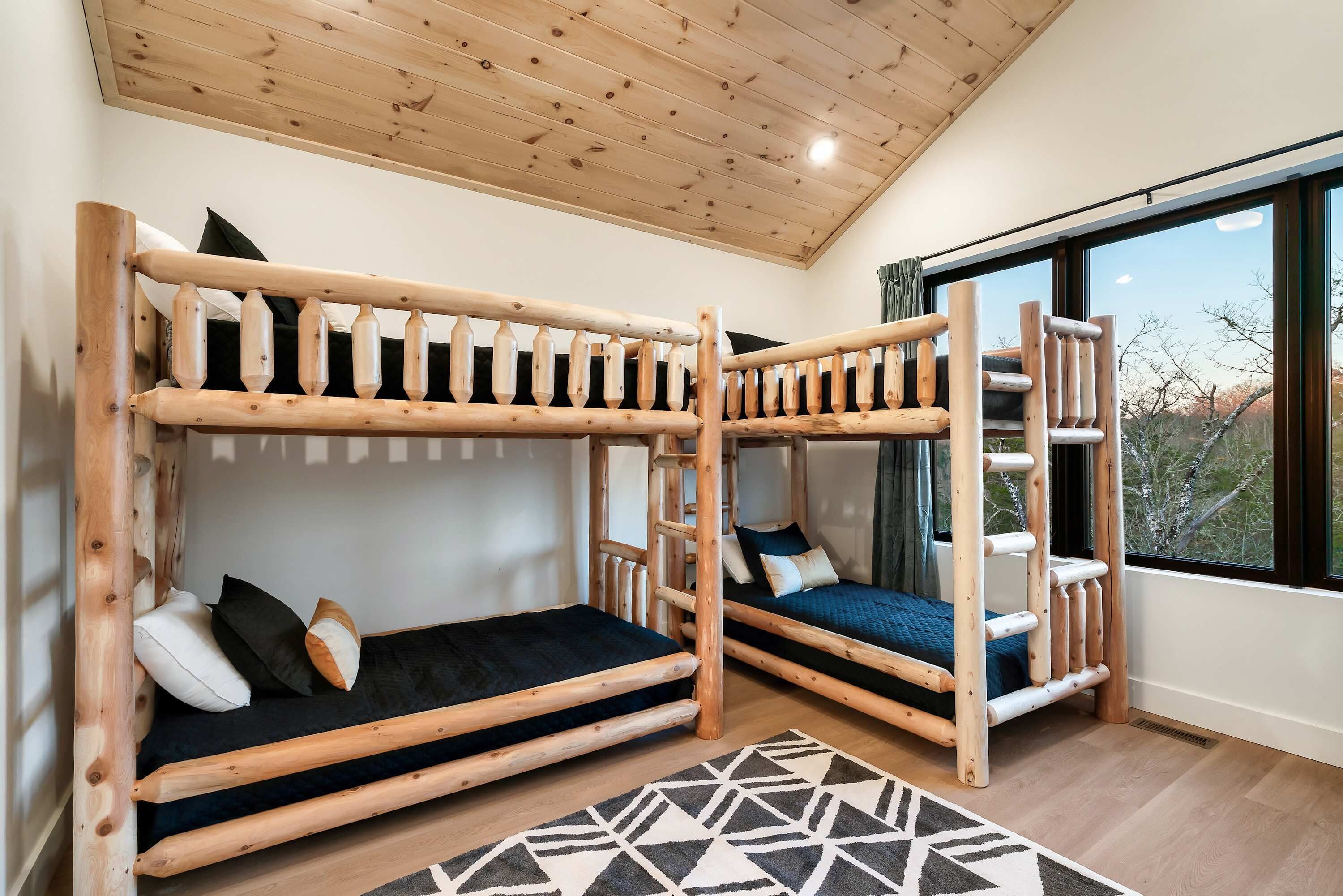Una habitación con dos literas de madera, un tapete con estampado geométrico y un amplio ventanal con vistas a árboles.