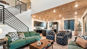 A modern living room with a green sofa, patterned armchairs, and a wooden coffee table. A staircase with black railings leads to an upper level.