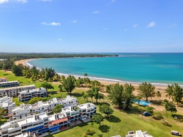 Un complejo turístico en la costa con varios edificios blancos, una playa y un mar azul cristalino.