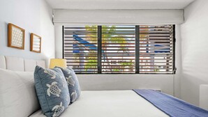 Un dormitorio con cama, almohadas, ventana con persianas y cuadros en la pared.