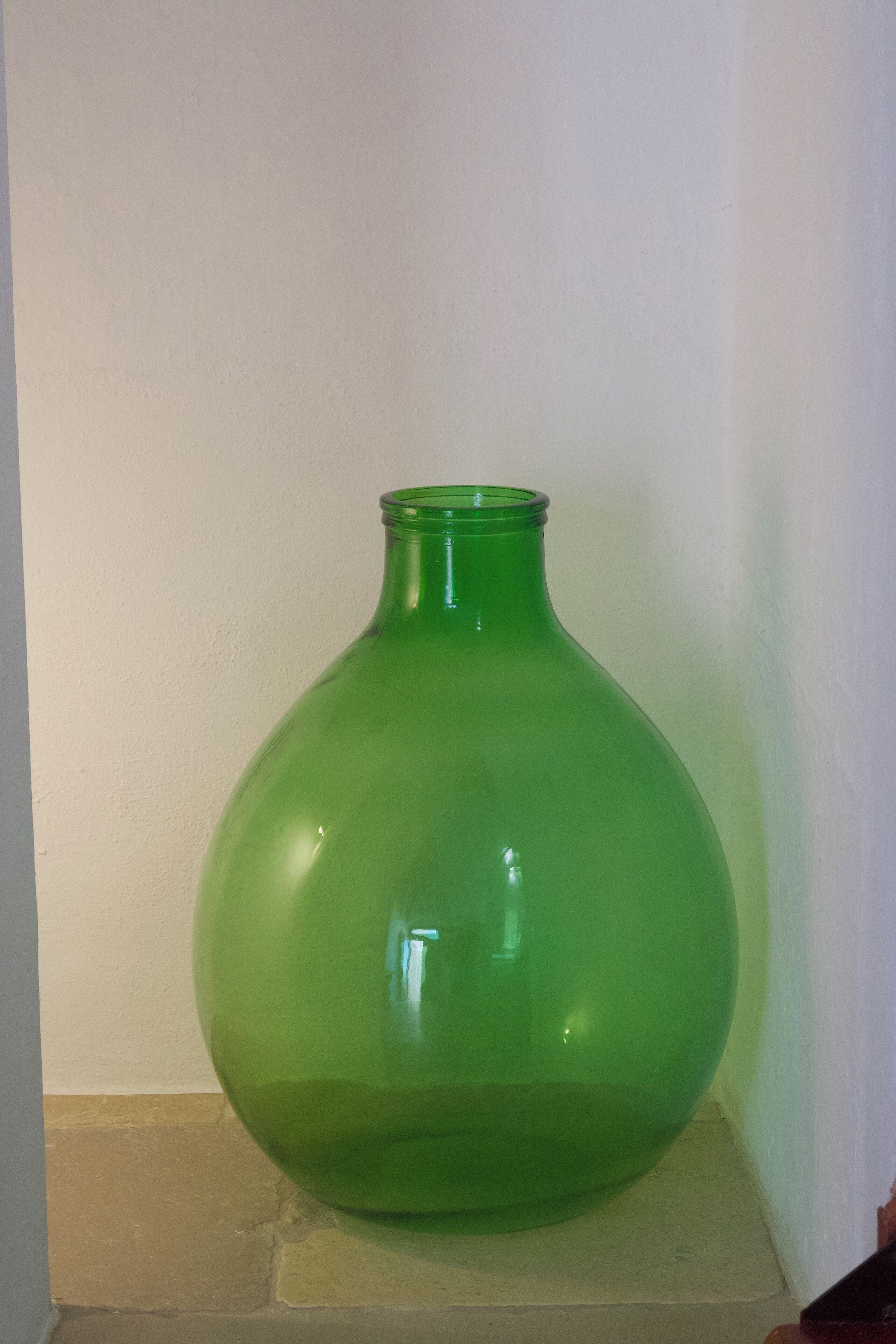 Un vase en verre vert, posé sur une étagère beige, devant un mur blanc.