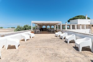Eine Terrasse im Freien mit weißen Plastikstühlen, einem überdachten Sitzbereich und Blick aufs Meer.