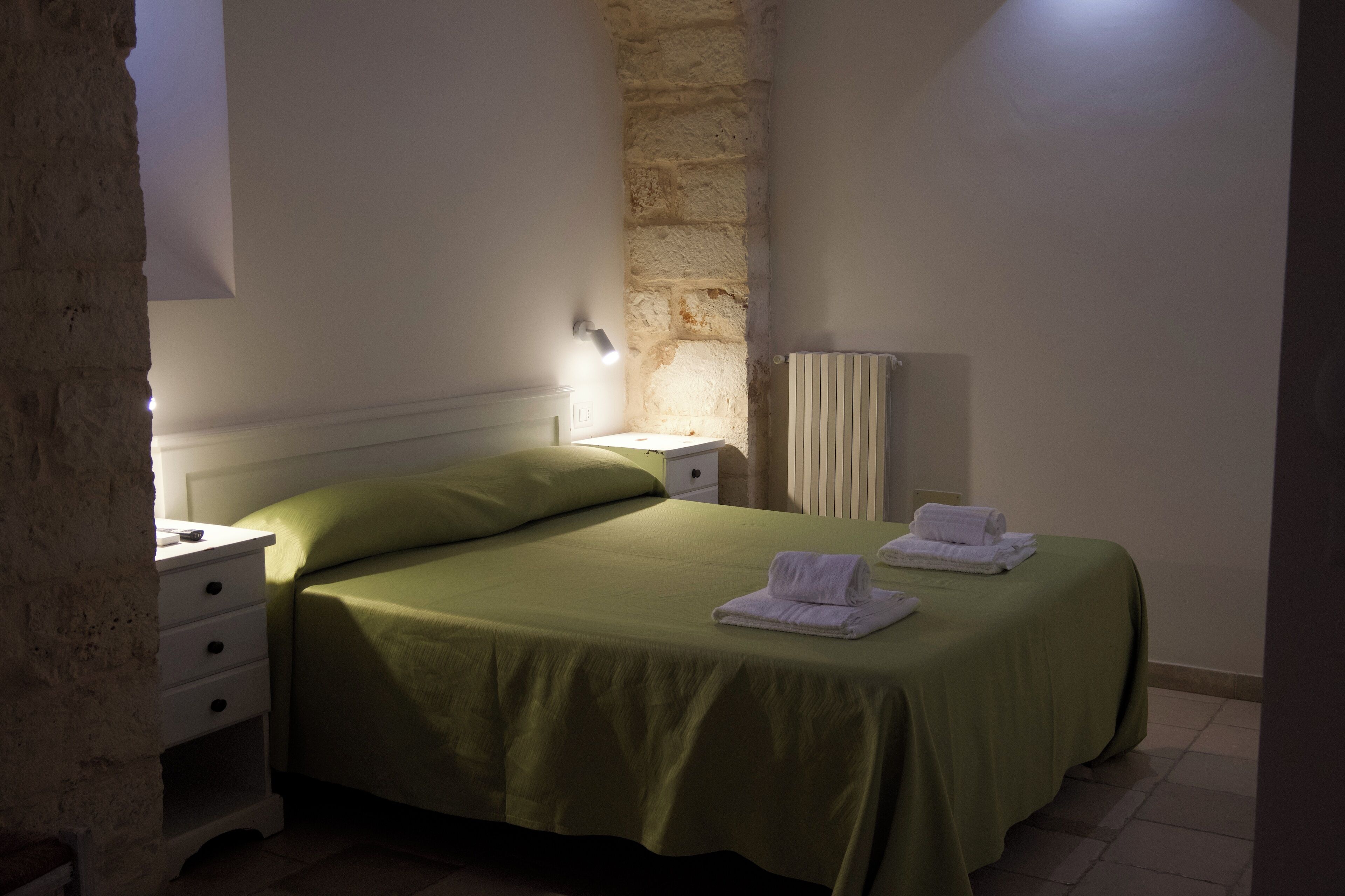 Einzimmerappartement mit einem Einzelbett, einer grünen Tagesdecke, weißen Handtüchern, einem Nachttisch, einer Lampe und einem Heizkörper.