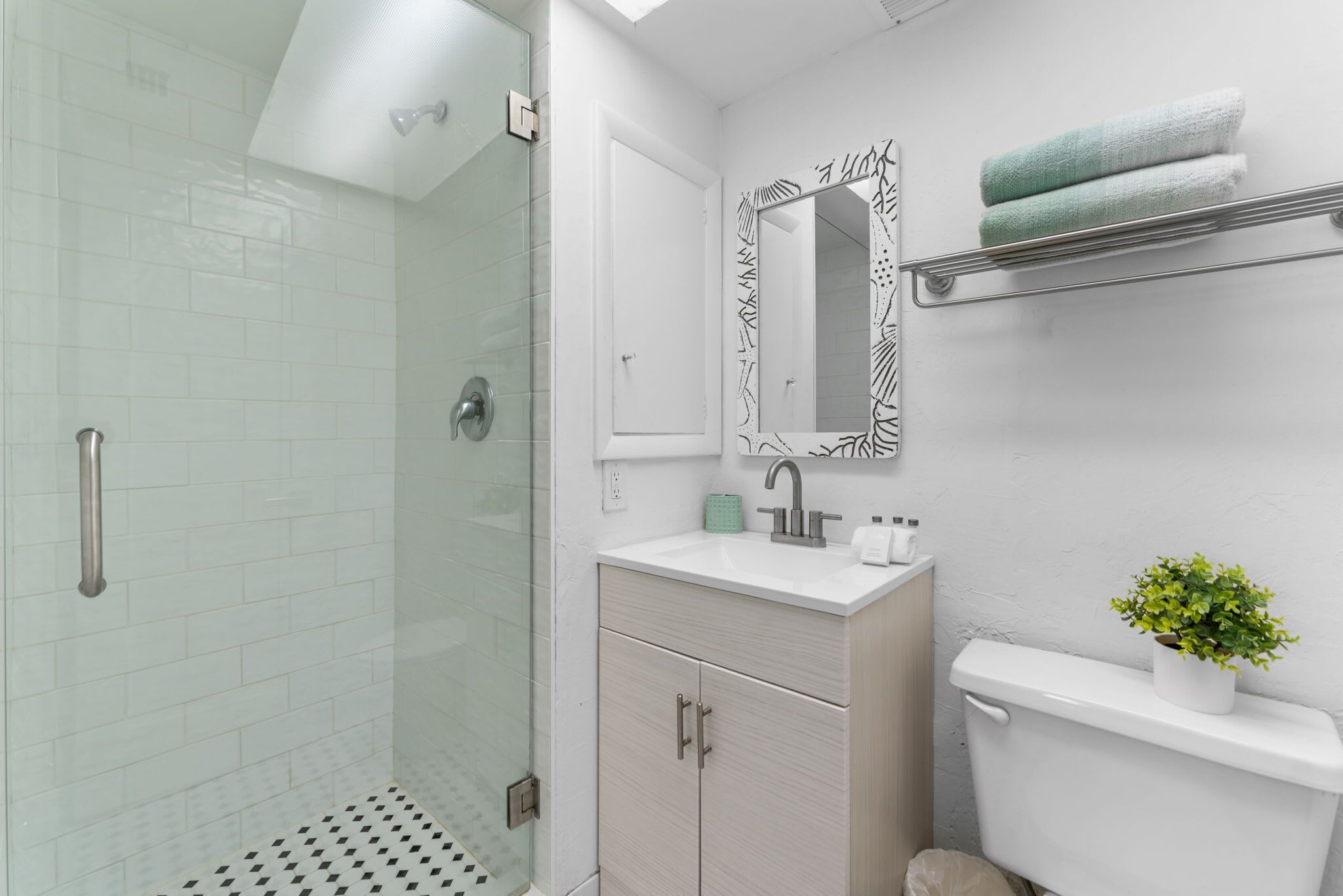 Un baño moderno con cabina de ducha de vidrio, un lavamanos blanco con espejo y un inodoro decorado con una planta.