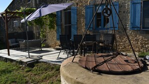 An outdoor seating area with wicker furniture and a swing seat, a stone building with blue shutters, and a pergola with a white umbrella.