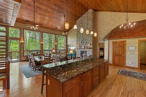A spacious kitchen with a large island, wooden cabinets, and a stone backsplash. There is a fireplace, large windows, and a dining area with a view of the outdoors.