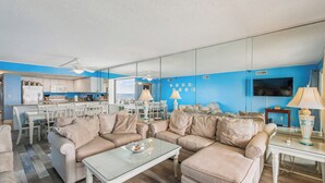 Een moderne woonkamer met beige banken, een glazen salontafel en een keuken op de achtergrond.