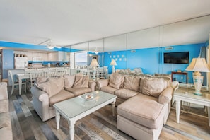 A modern living room with beige sofas, a glass coffee table, and a kitchen in the background.