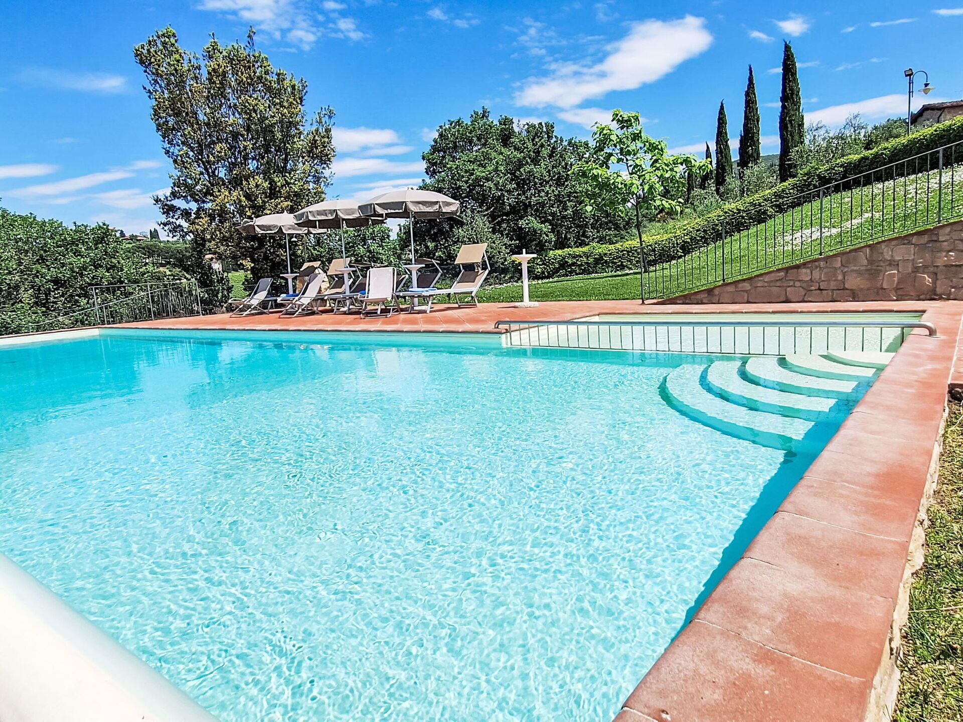 A clear swimming pool with lounge chairs and umbrellas, surrounded by greenery and stone walls.