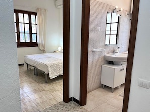 Un dormitorio con cama, mesita de noche, ventana con cortinas y baño con lavamanos y espejo.
