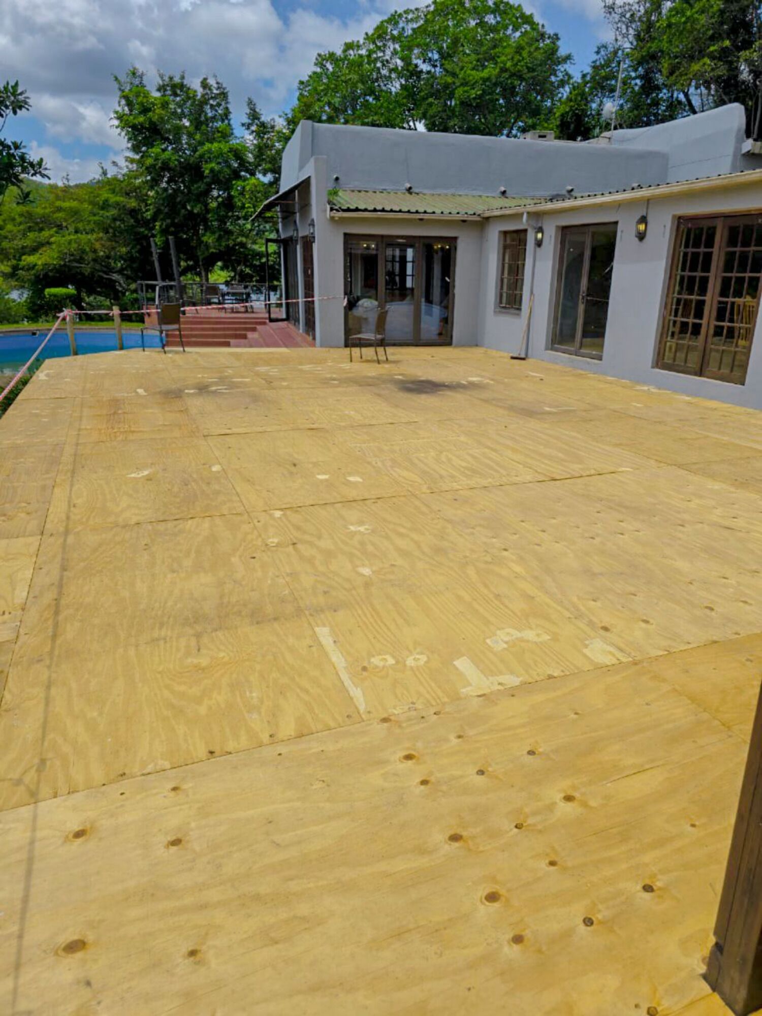 Une terrasse en bois avec une piscine, un espace salon couvert et un bâtiment doté de grandes fenêtres et d’une porte.