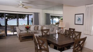 Un comedor con mesa y sillas de madera, un sofá y vistas a la playa a través de amplios ventanales.