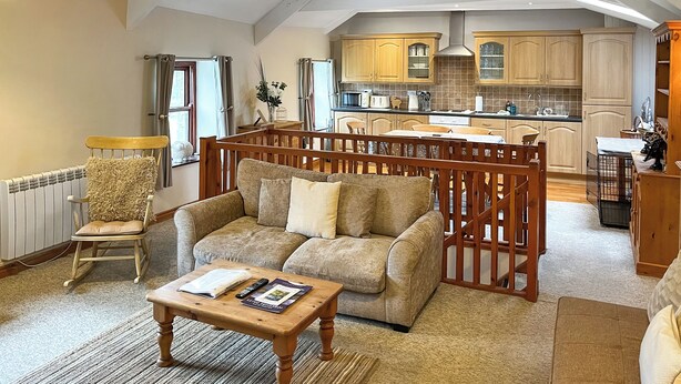 A cozy living room with a sofa, a rocking chair, and a coffee table. A kitchen is visible in the background with wooden cabinets and a dining area. - 4 Swallowholm Cottage (Arkengarthdale, near Reeth)