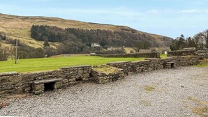 A stone wall with a small opening, a grassy field, and a hill with trees in the background. - 4 Swallowholm Cottage (Arkengarthdale, near Reeth)