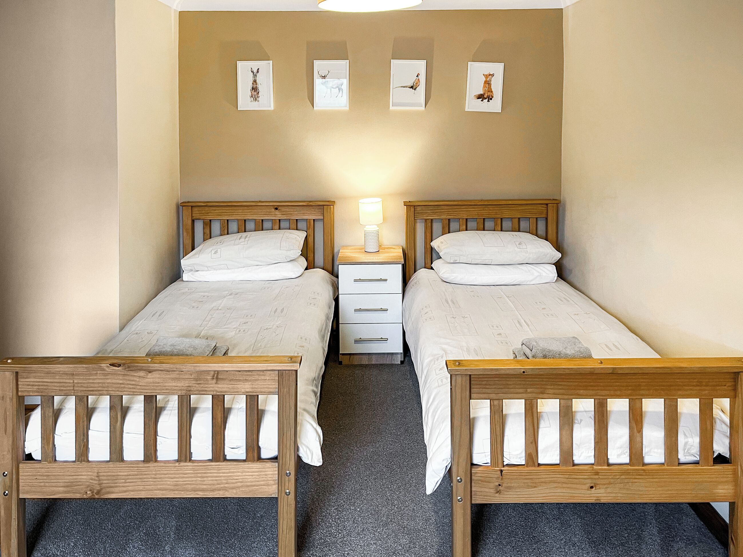 Una habitación doble con dos camas de madera, una mesita de noche con lámpara y tres cuadros enmarcados en la pared.