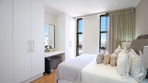 Un dormitorio moderno con una cama grande, un armario empotrado, un escritorio pequeño y vistas a la ciudad a través de amplios ventanales.