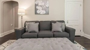 Ein Wohnzimmer mit grauem Sofa, einem раnschen Ottomanen, einer Stehlampe und einem gerahmten abstrakten Kunstwerk an der Wand.