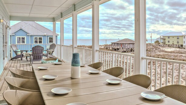 Un área de comedor al aire libre, techada, con vistas a la playa y una casa azul al fondo.