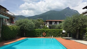 A rectangular swimming pool with a clear blue water surface, surrounded by a red-tiled deck. In the background, there are buildings with balconies and a mountain range. - Peler House at Benaco - Style, Lake view & Pool (Toscolano Maderno)
