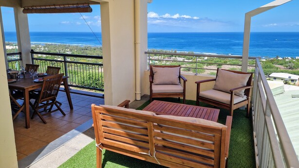 A balcony with a wooden table and chairs, a cushioned bench, and a view of the ocean. - Large T3 • Breathtaking ocean view - Saint-Gilles (Saint-Paul)