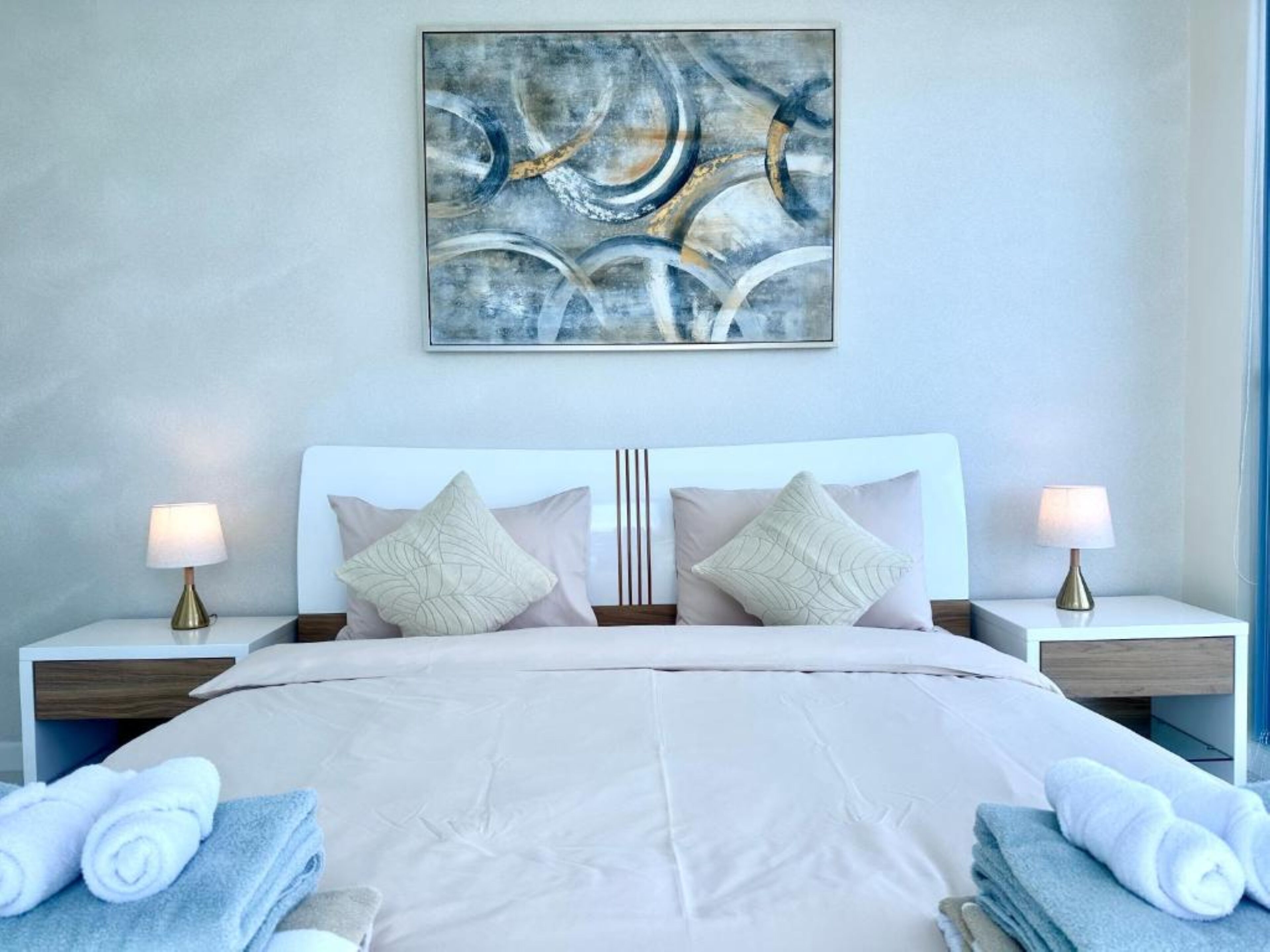 Une chambre à coucher moderne avec un grand lit, deux tables de chevet avec des lampes et un tableau abstrait accroché au mur.