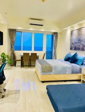 A bedroom with a bed, a desk, and a chair. There is a window with curtains, a plant, and a painting on the wall. - La Mirada Deluxe Suites with Oceanview (Lapu-Lapu)