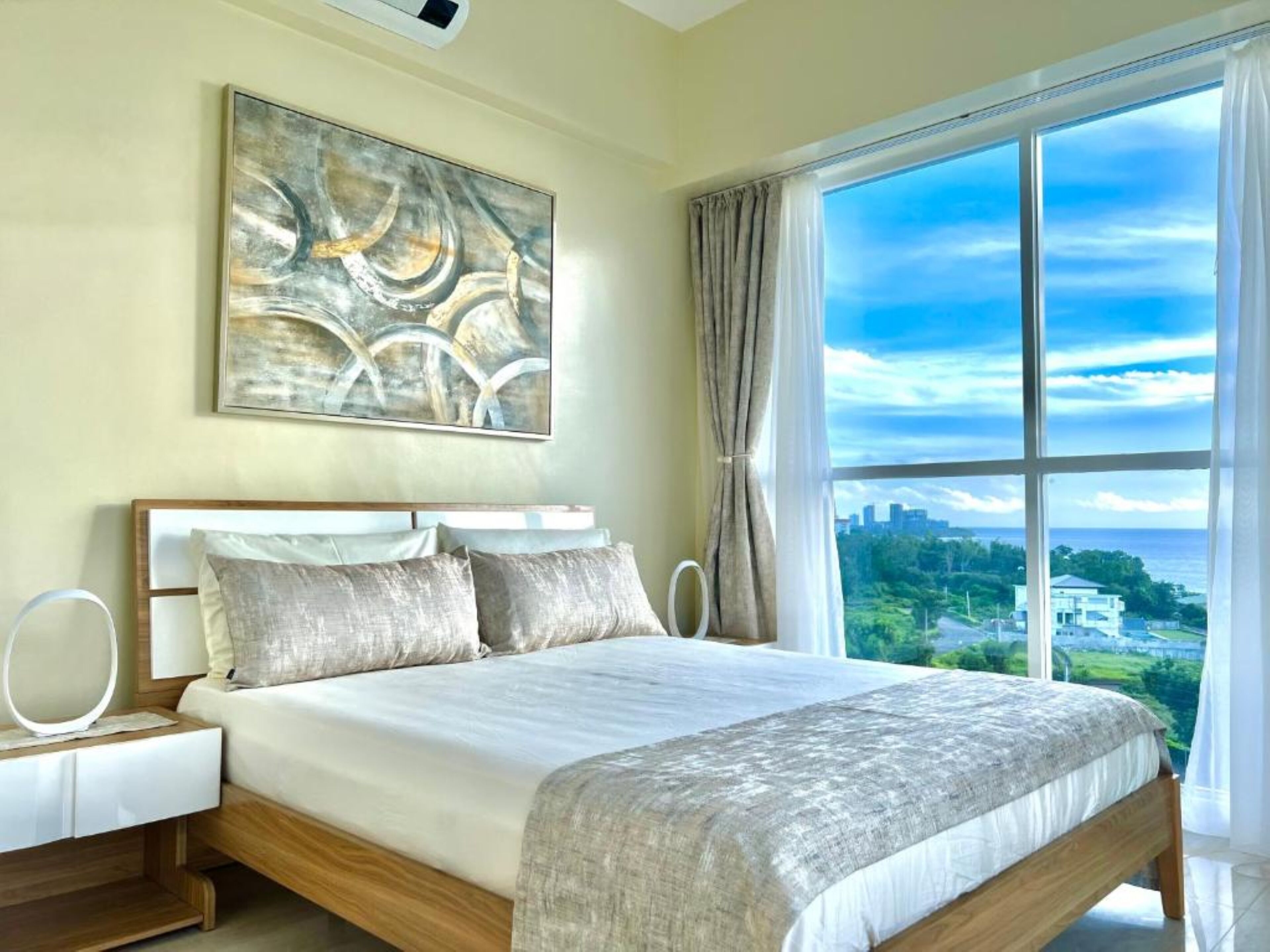 A bedroom with a large bed, a bedside table, a wall-mounted artwork, and a view of a cityscape through a large window.