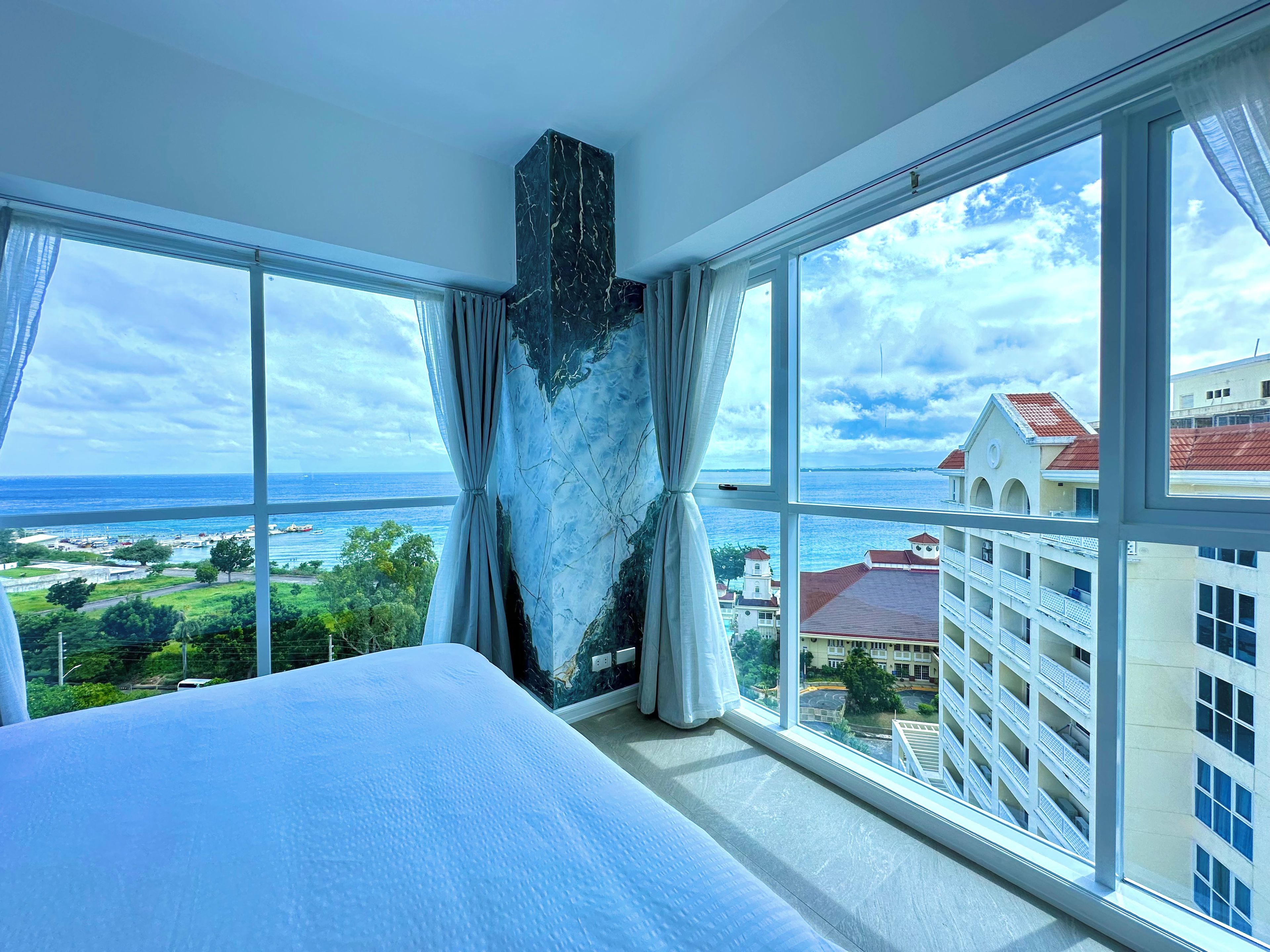 A hotel room with a large bed, a marble accent wall, and a view of the ocean and buildings.