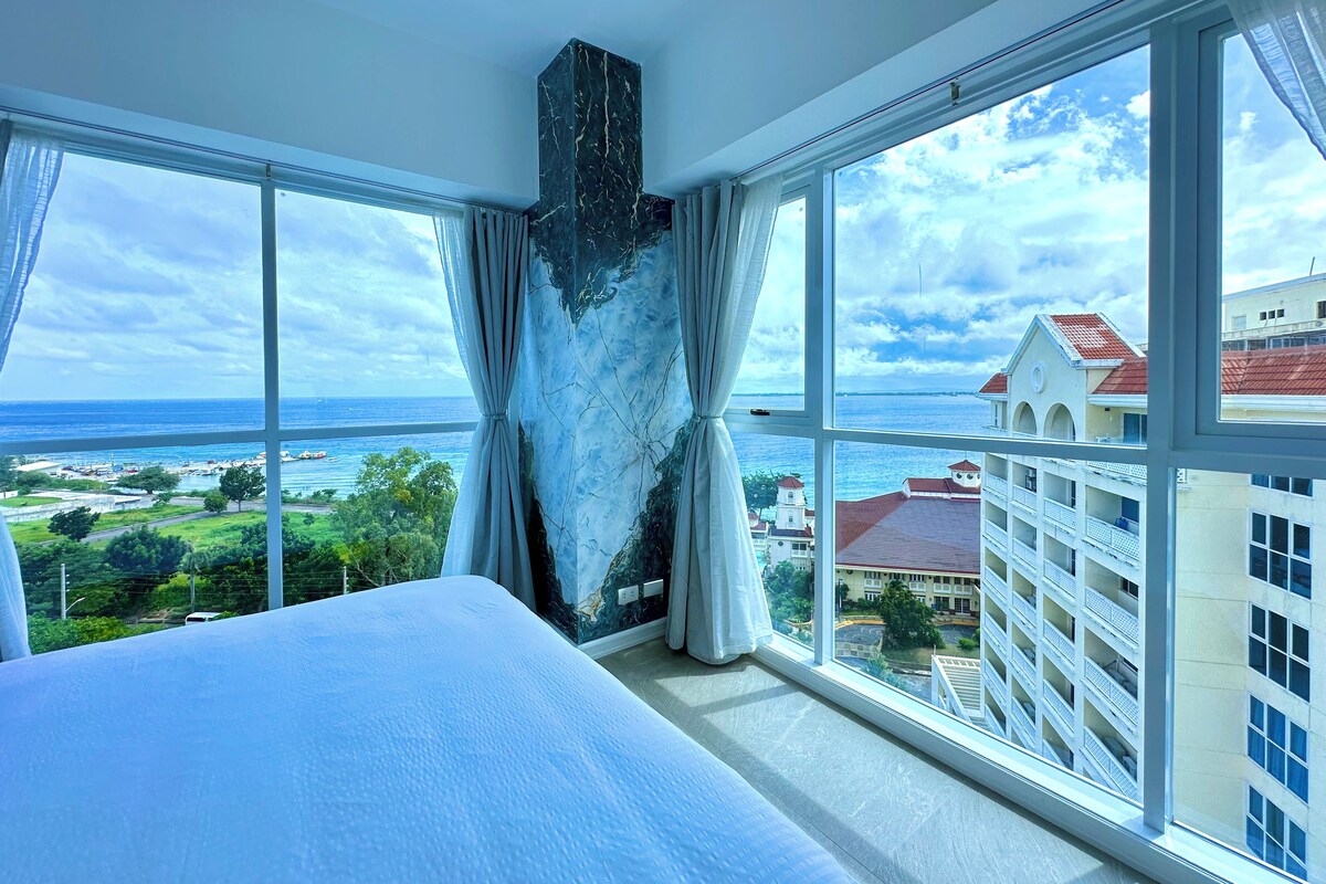 Une chambre d’hôtel dotée d’un grand lit, d’un mur décoratif en marbre et offrant une vue sur l’océan et les bâtiments.
