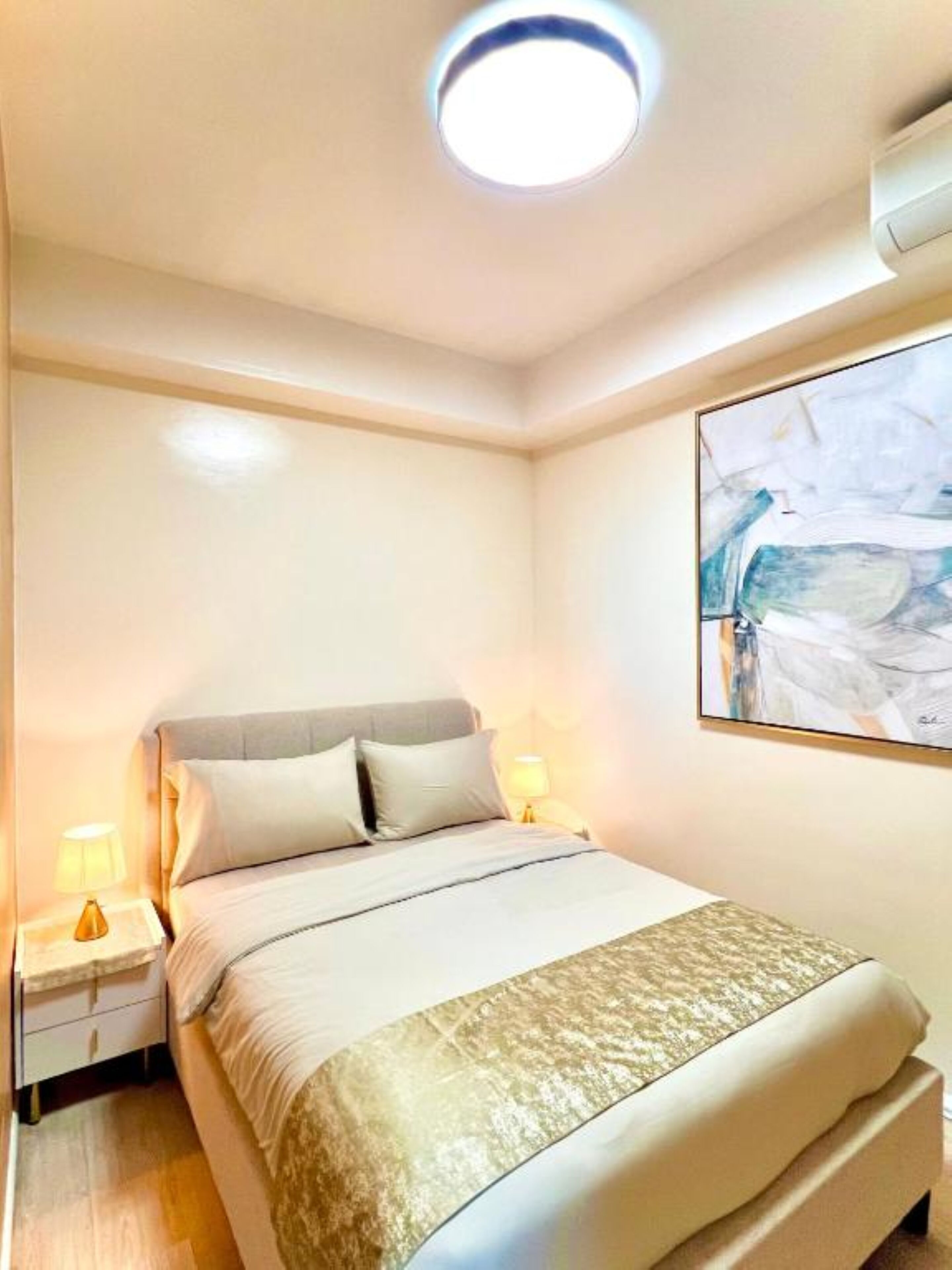 Una cama bien hecha con cabecera, dos mesitas de noche con lámparas y un cuadro abstracto grande en la pared.