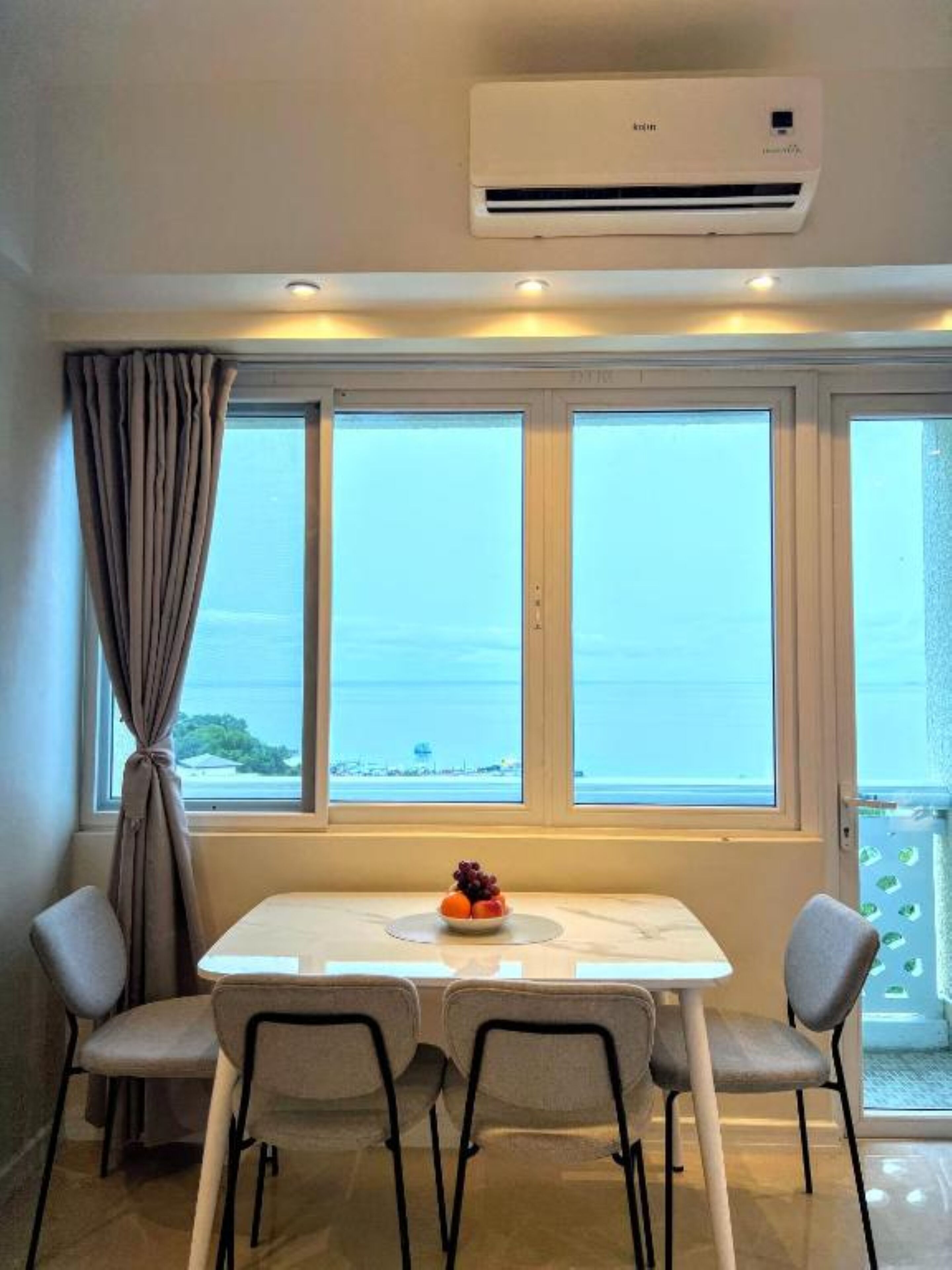 A room with a table and chairs, a window with a view of the sea, and an air conditioning unit above the window.