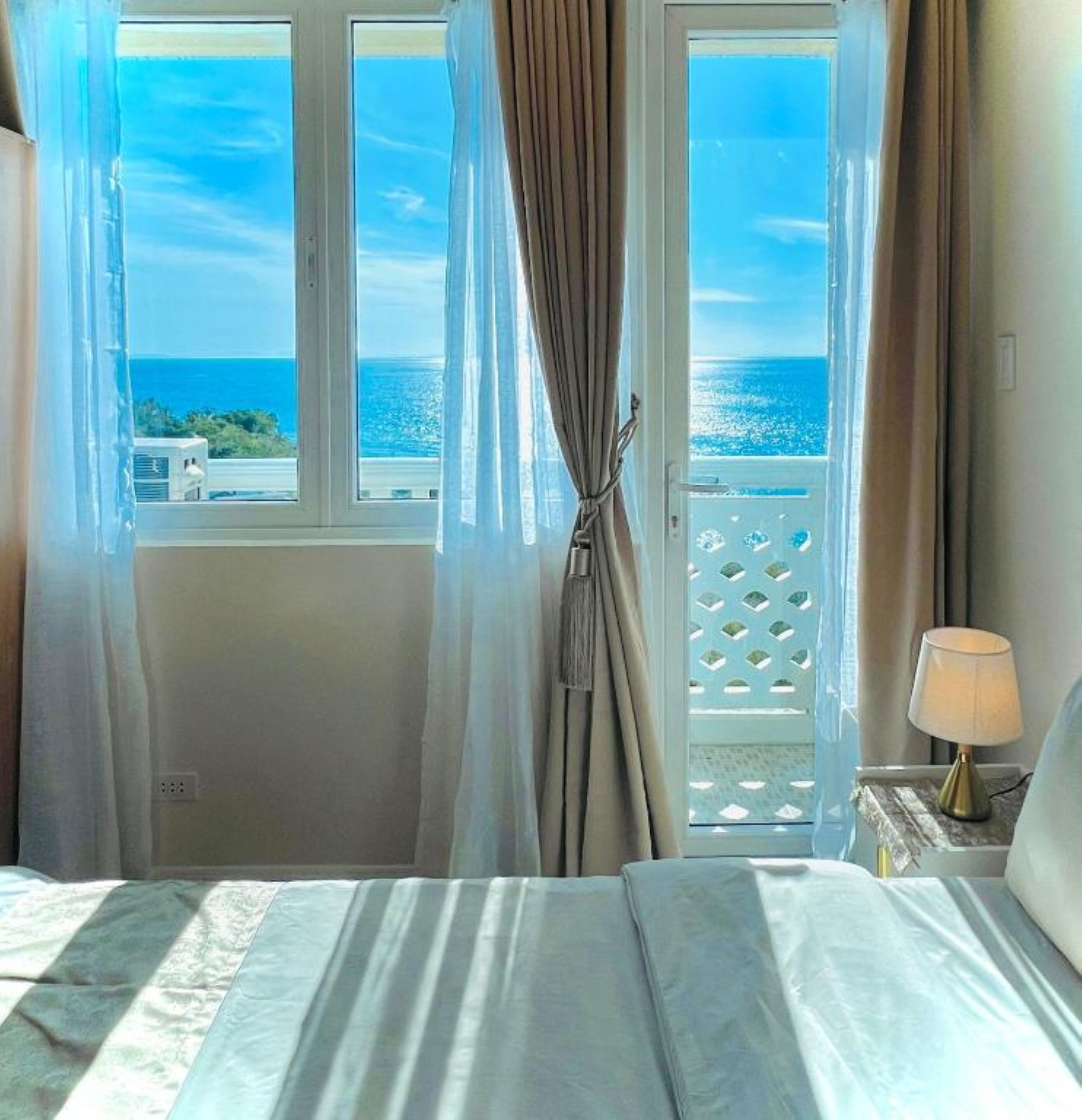 Ein Hotelzimmer mit Bett, Nachttisch mit Lampe und Blick auf das Meer durch ein Fenster mit Vorhängen.