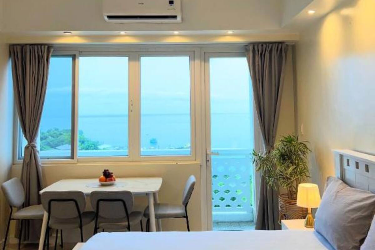 A hotel room with a bed, a dining table with chairs, a small table with fruit, and a view of the sea through large windows.