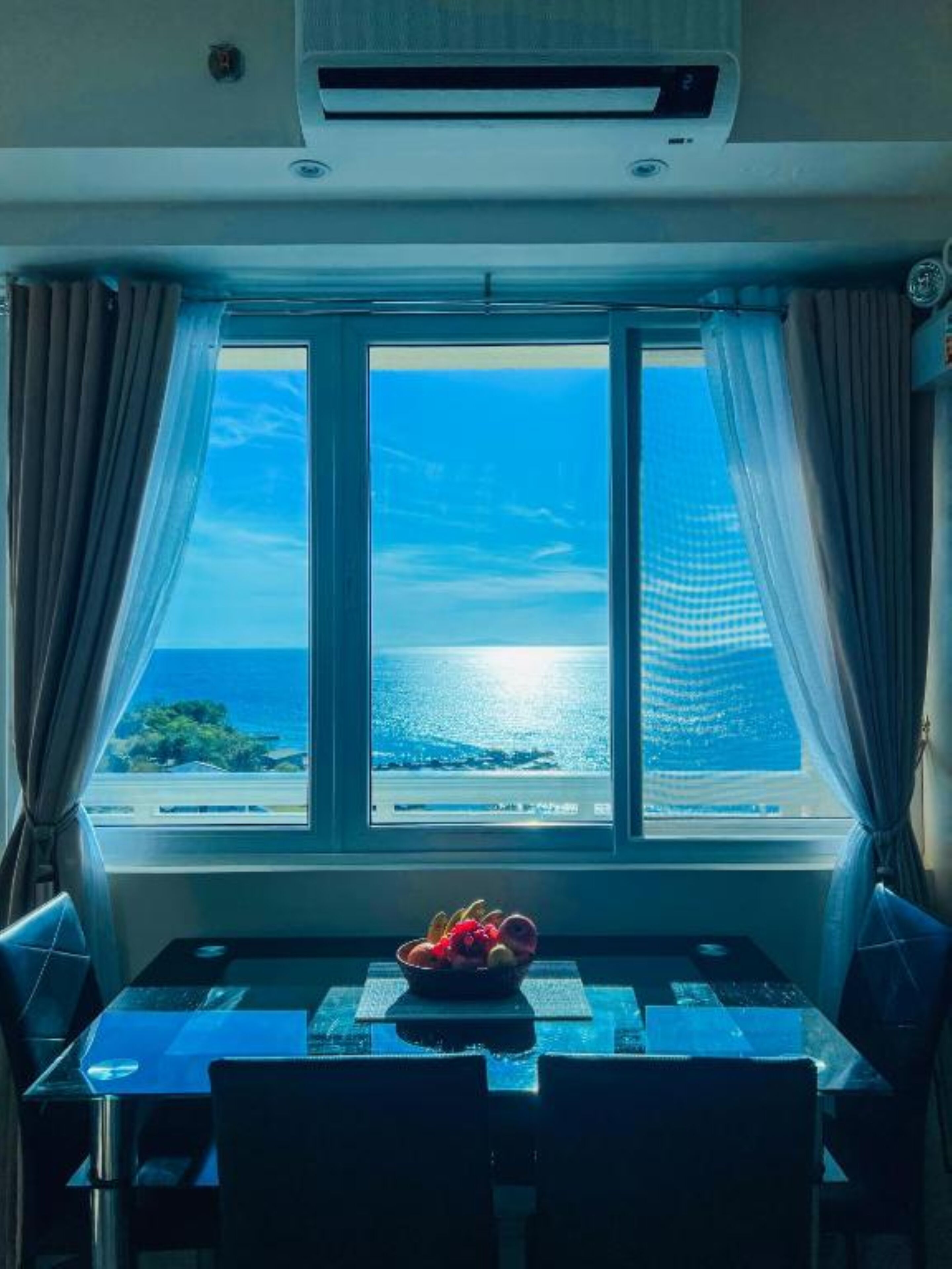 Ein Zimmer mit Tisch und Stühlen, einem Fenster mit Vorhängen und Blick aufs Meer.