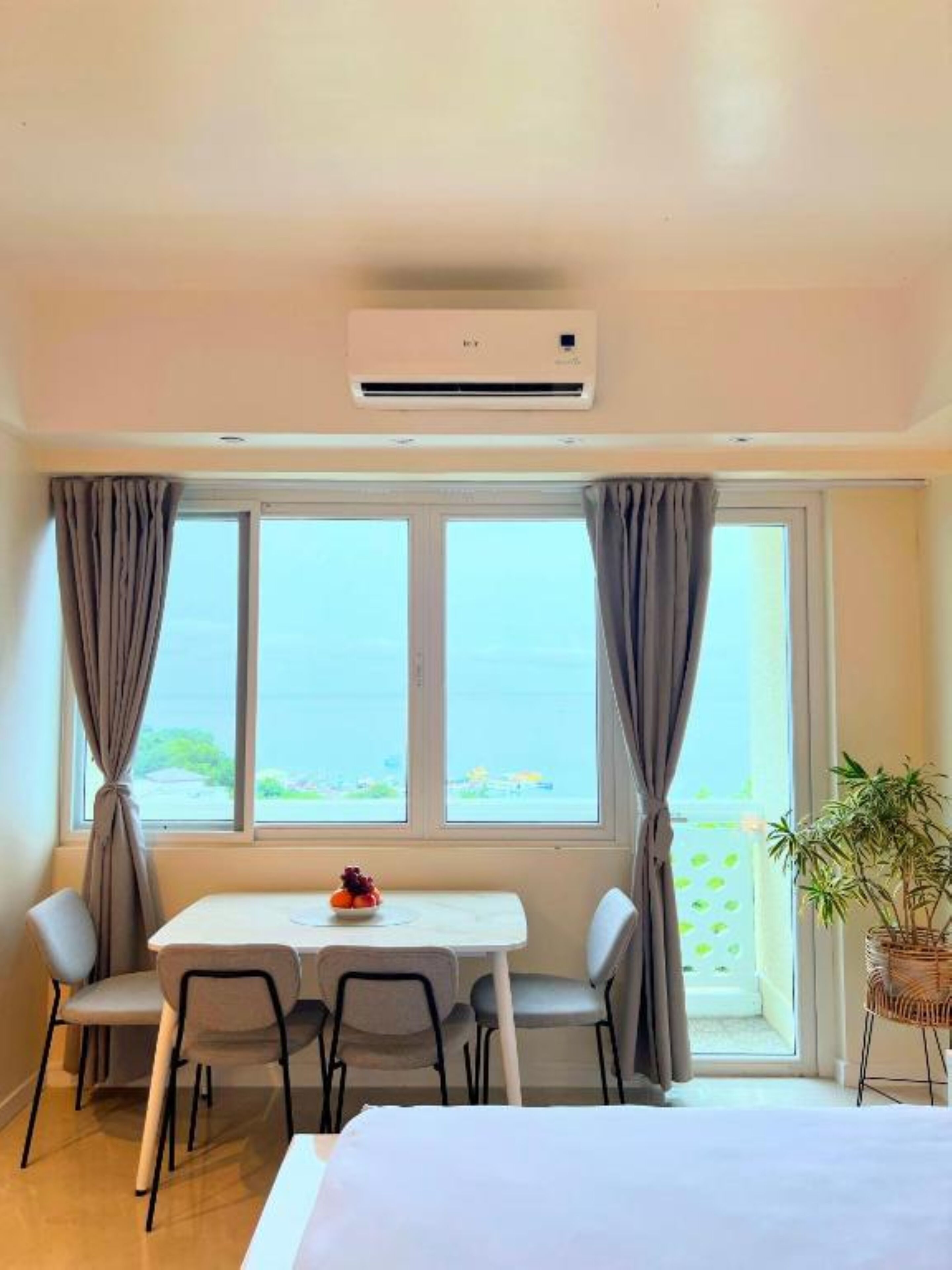 A hotel room with a bed, a dining table with chairs, a potted plant, and a view of the sea through large windows.