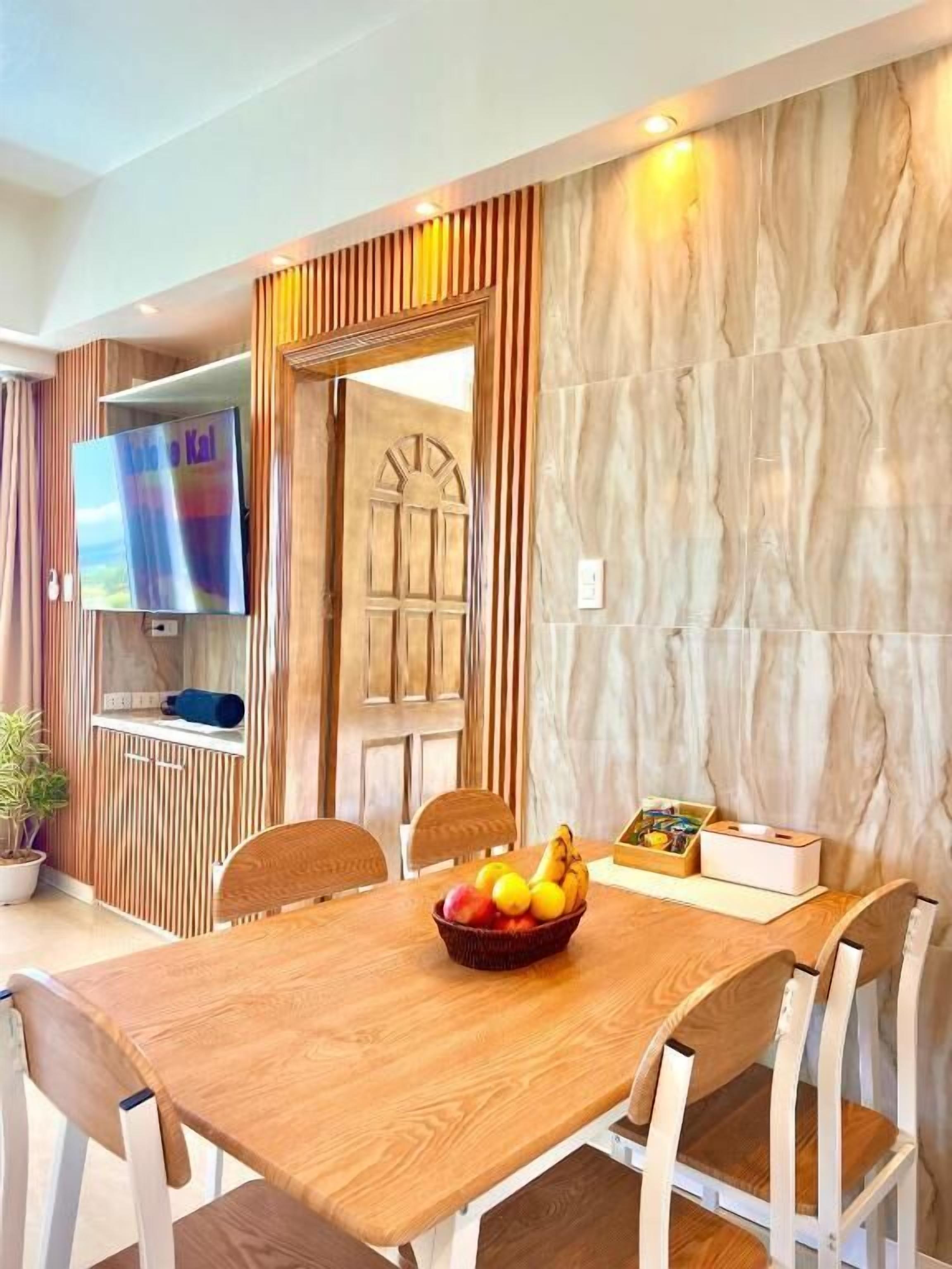 A dining area with a wooden table, chairs, a fruit bowl, and a television on a wall.