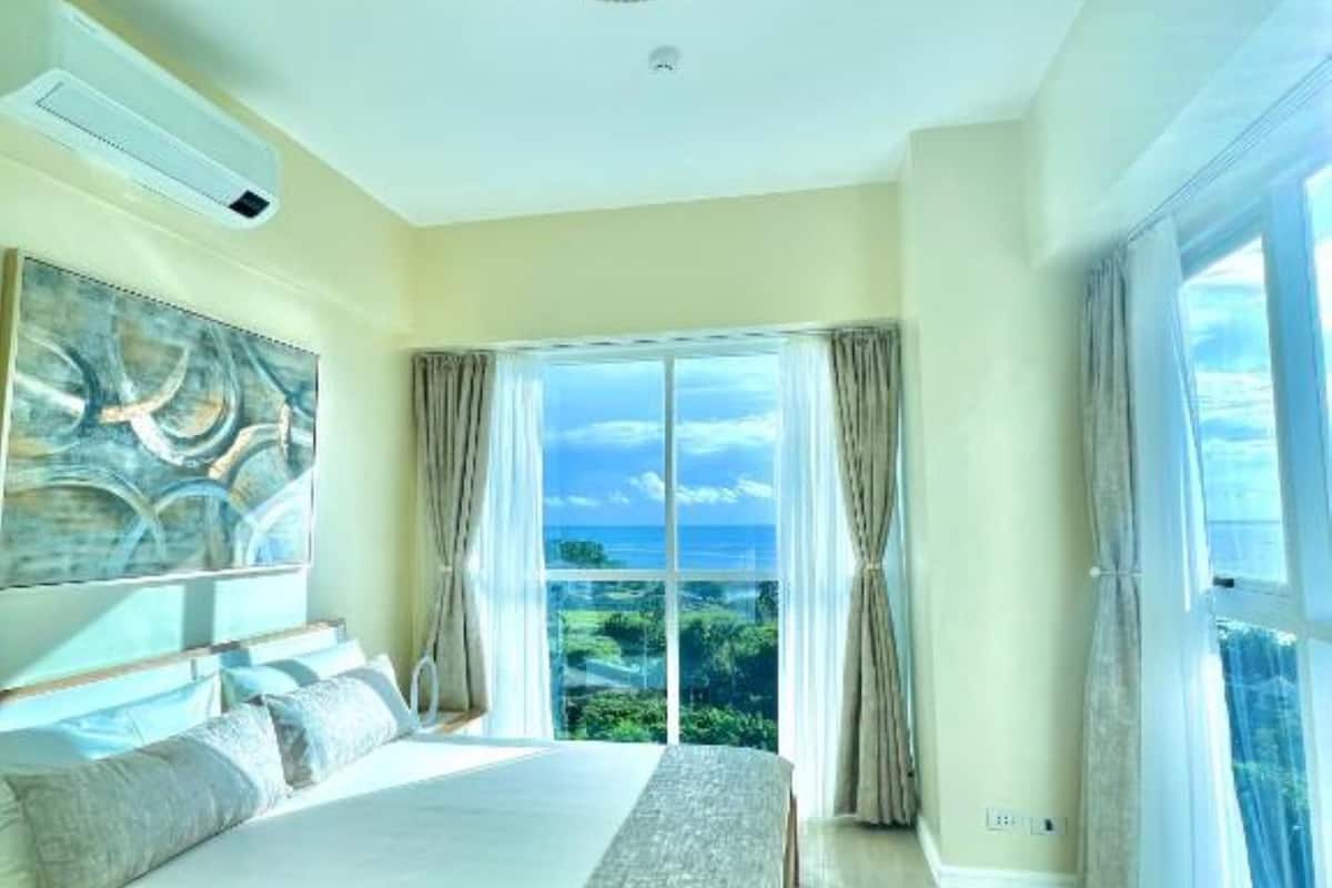 A spacious bedroom with a large bed, a ceiling light fixture, and a view of the outdoors through a large window.