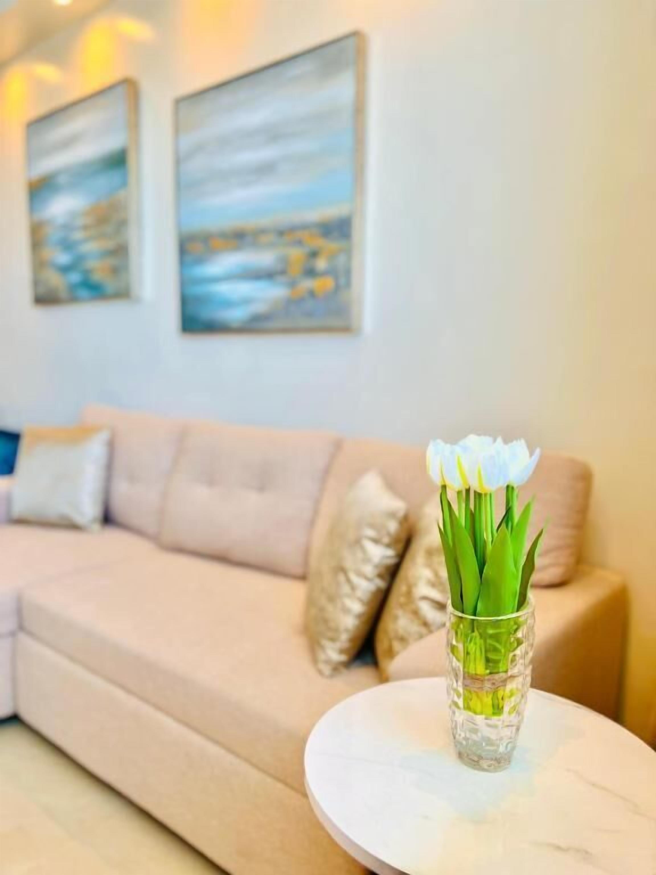 A living room with a beige sofa, a glass vase with white flowers on a round table, and two landscape paintings on the wall.