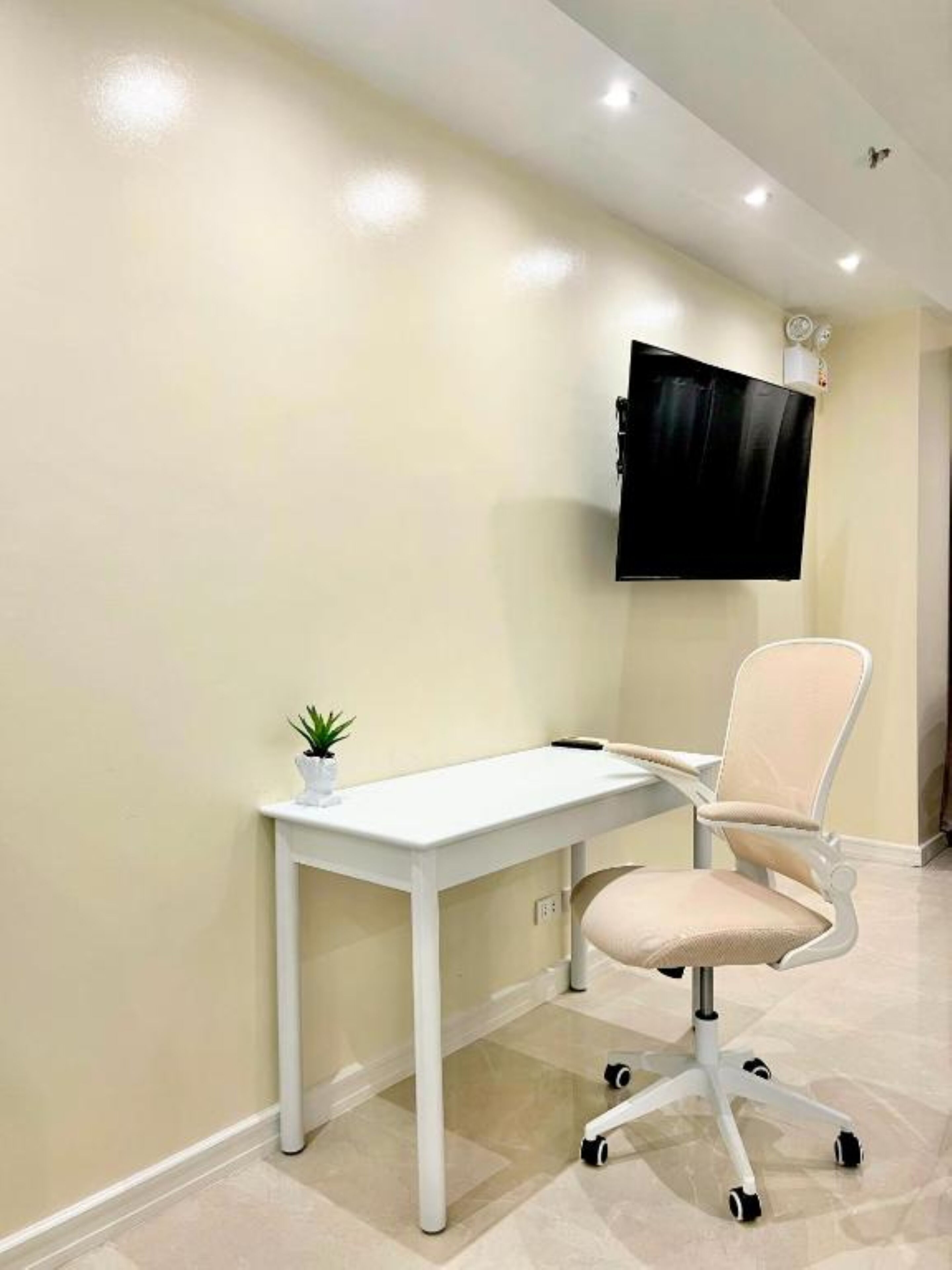 A modern office space with a white desk, a cushioned office chair, a small potted plant, and a mounted flat-screen TV.