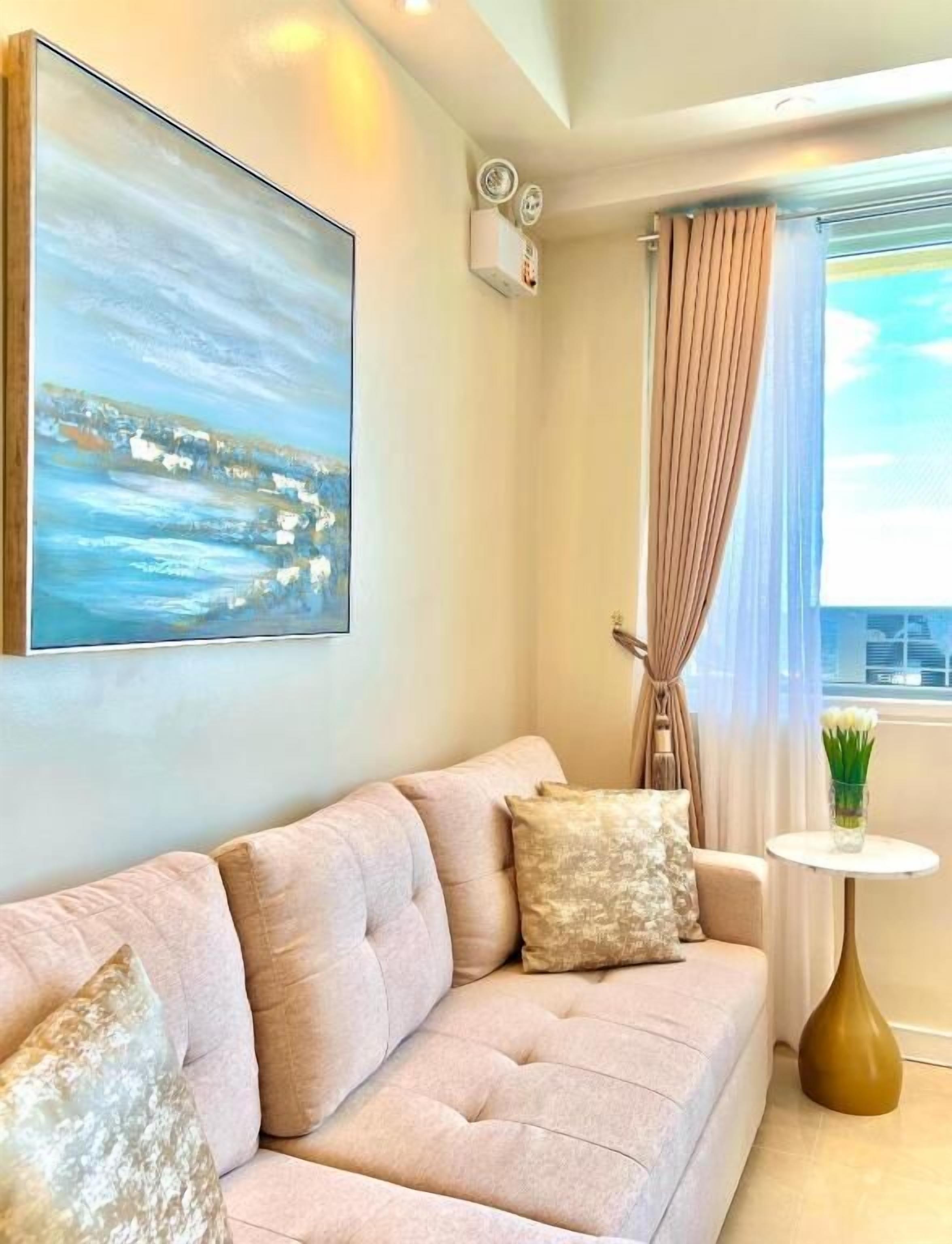 Un salon moderne avec un canapé beige, un tableau représentant un paysage côtier, une petite table ronde ornée d’un vase, et une vue sur la ville à travers une fenêtre avec des rideaux.