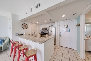 A modern kitchen with a central island, red bar stools, stainless steel appliances, and a light blue accent wall. - Beachfront Long Beach Tower IV 1205, Pools, Hot Tub, FREE Golf & Perks! (Panama City Beach)