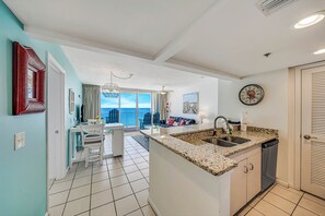 A modern kitchen with a granite countertop, a dishwasher, and a dining area with a white table and chairs. - Beachfront Long Beach Tower IV 1205, Pools, Hot Tub, FREE Golf & Perks! (Panama City Beach)