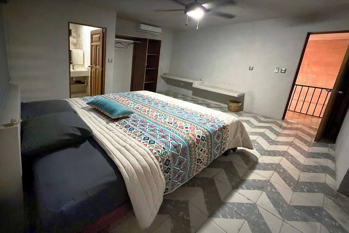 A bedroom with a bed, a ceiling fan, a bathroom with a mirror, and a tiled floor.