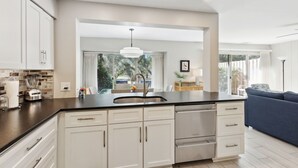 Een moderne keuken met witte kastjes, een zwart aanrecht en een inbouwspoelbak. Er is een zithoek met een blauwe bank en uitzicht op de omgeving.