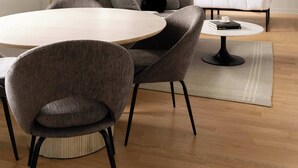 A modern living room with a round dining table, two grey armchairs, a sofa, a floor lamp, and abstract wall art.
