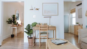 A modern living room with a wooden desk, a comfortable sofa, and a coffee table. There is a large plant and a bookshelf with decorative items. - Walk to Dining, Beach & Park~Family Friendly Home! (Cape Charles)